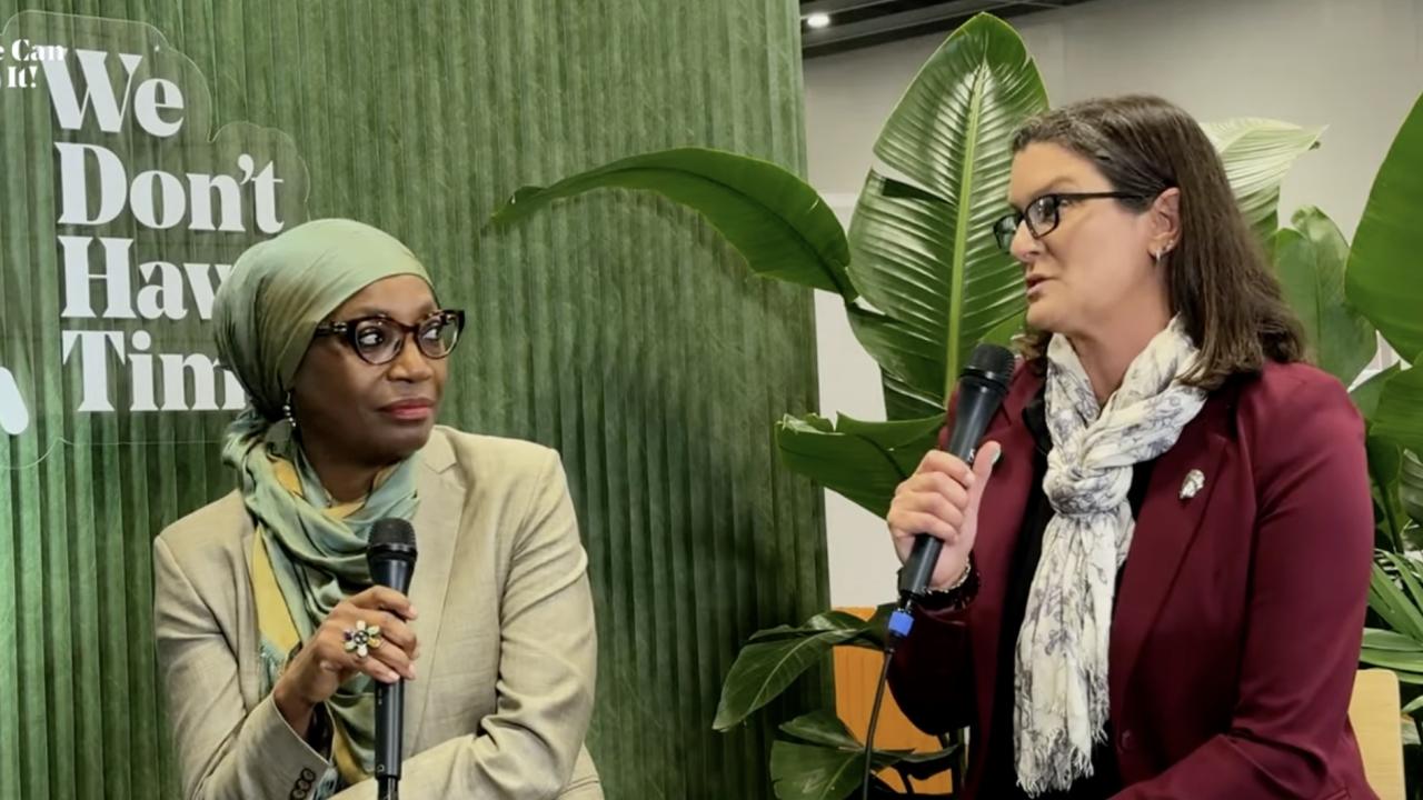 Fatima Denton,left, hold microphone and turns head to Tessa Hill, who is speaking into microphone. A green background and plant are behind them with the words We Don't Have Time printed on backdrop.