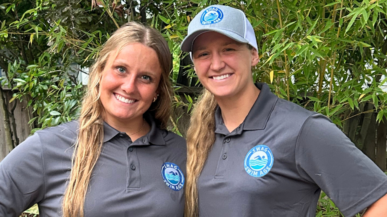 Two smiling women stand together outdoors, wearing matching gray polo shirts and caps.