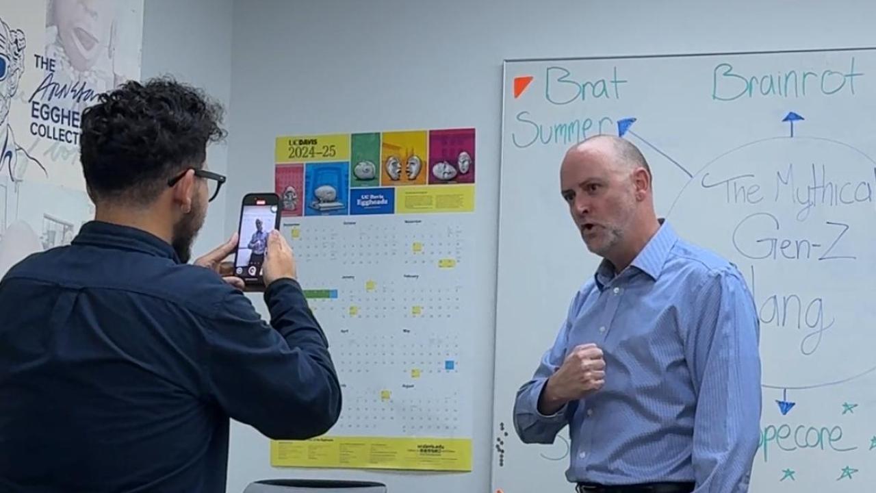 A person films UC Davis Undergraduate Admissions Executive Director Robert Penman with a smartphone as he poses in front of a whiteboard labeled “The Mythical Gen Z Slang” inside a conference room.