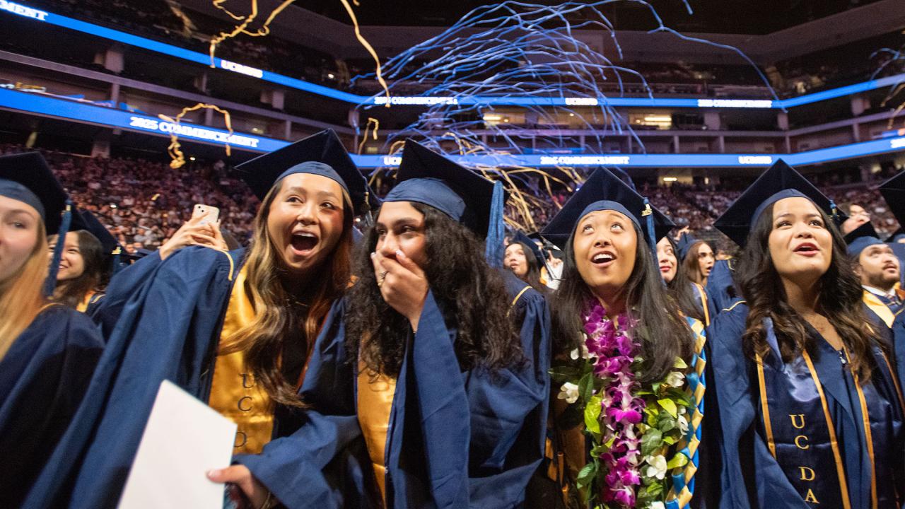 Students in graduation caps and gowns react in surprise and joy and streamers fall from above at an undergraduate commencement at the Golden 1 Center in June 2025. 