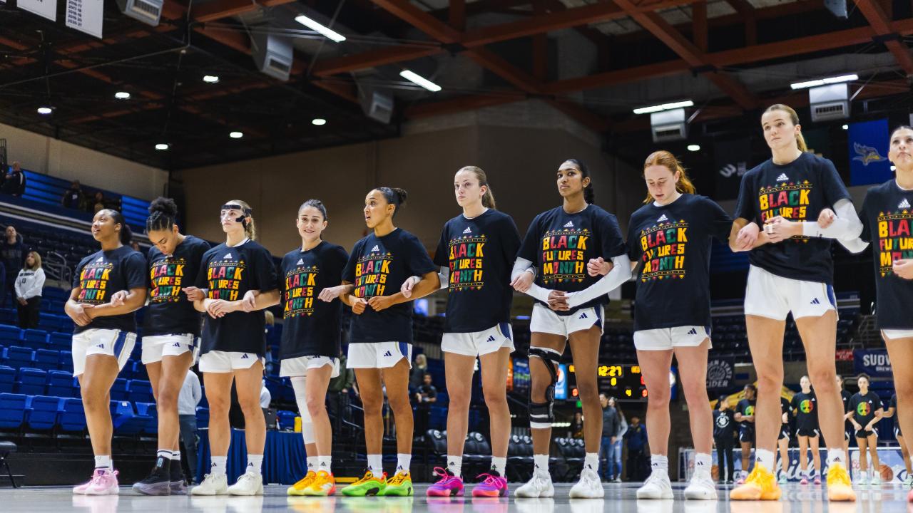 Inside a basketball gymnasium, a row of women basketball players face the camera. They're all wearing t-shirts that read 'Black Futures Month'.