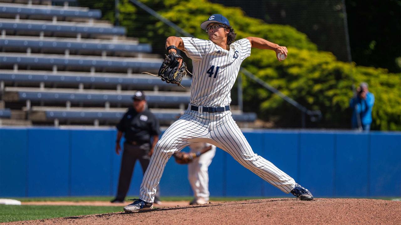 A baseball pitcher in mid-pitch, about to release the ball. He is wearing a white uniform with blue pinstripes, glasses and a blue hat.