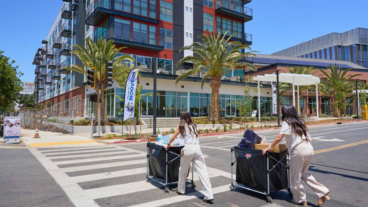 Two people push moving carts across a street toward housing at Aggie Square