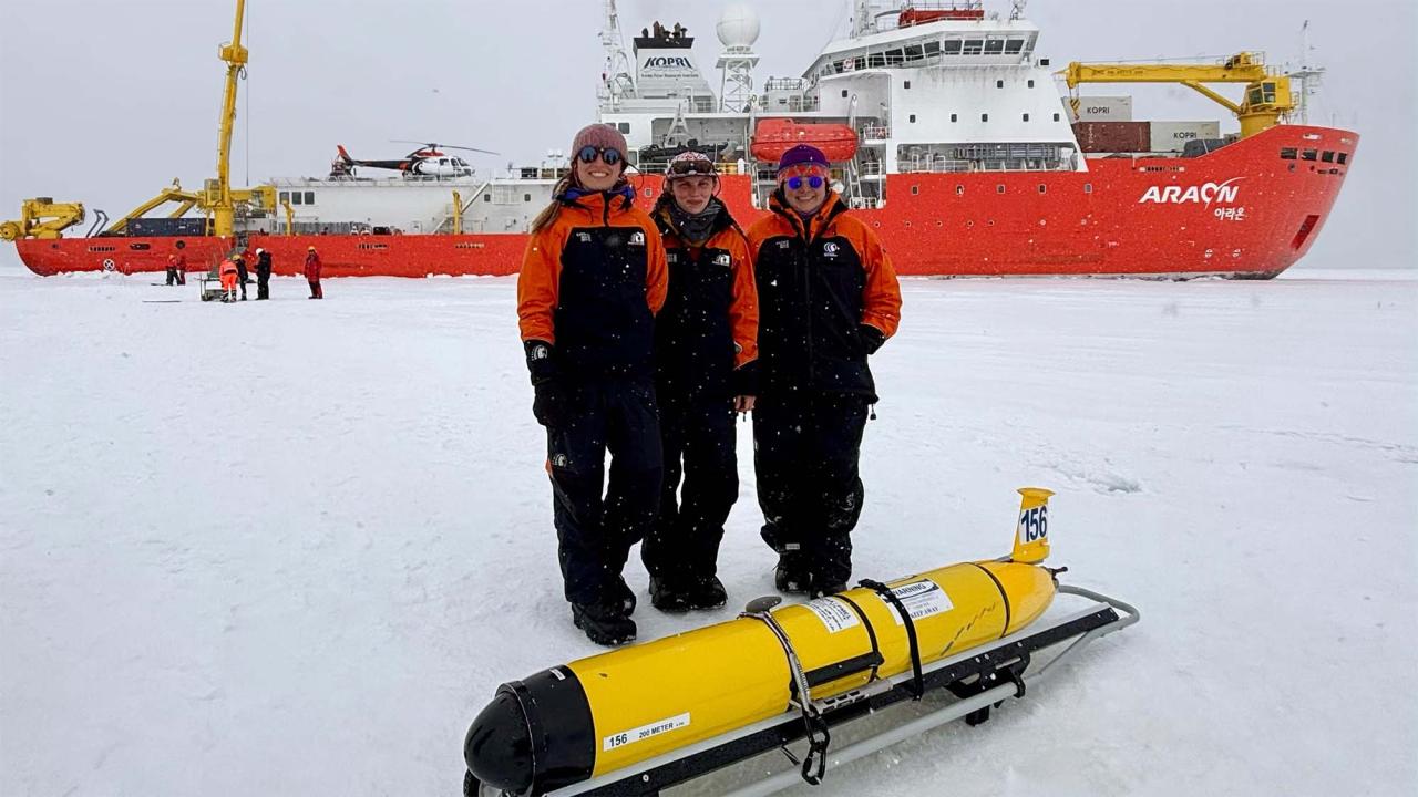 Three scientists in orange jackets stand on snow with a yellow underwater drone and a research ship behind them.