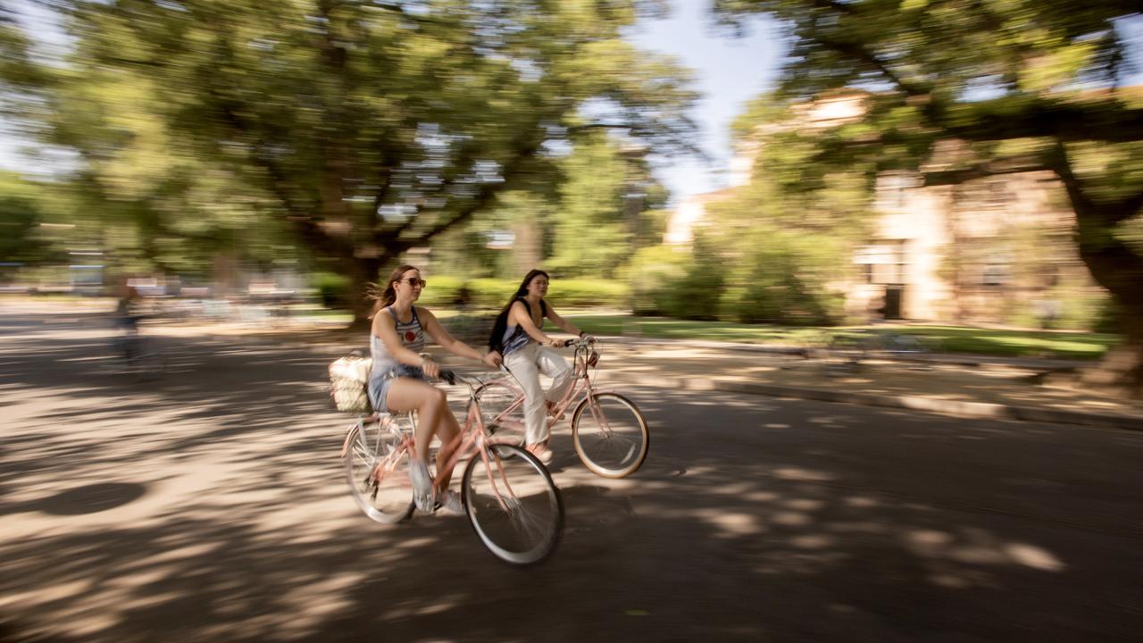 Two students bike along a campus road against a blurred background of trees