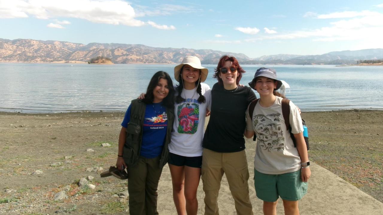 Four smiling people stand side by side on a lakeshore with their arms around each other. They are dressed in casual outdoor clothing and hats, with a scenic lake and mountain range in the background under a blue sky with scattered clouds.