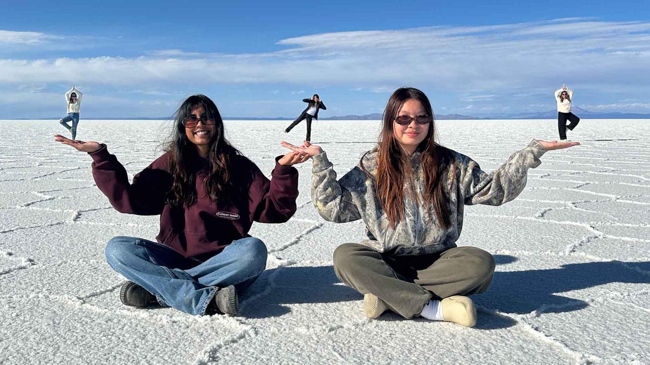 "On a sunlit salt flat, two women wearing sunglasses sit on the ground, playfully holding up smaller figures in their hands through forced perspective. The smaller figures in the background stand in yoga poses, with the salt flats stretching out beneath a wide, clear sky."