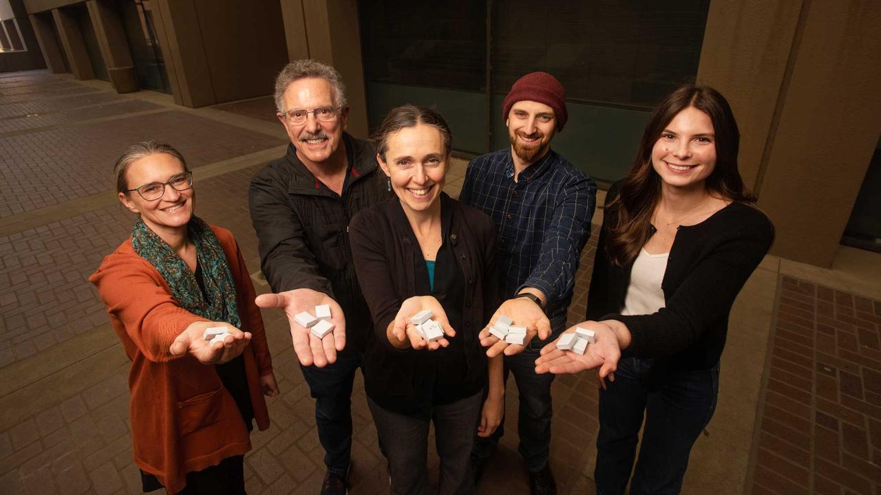 Portrait of five people standing in a group, each holding out a hand with white cubes toward the camera.