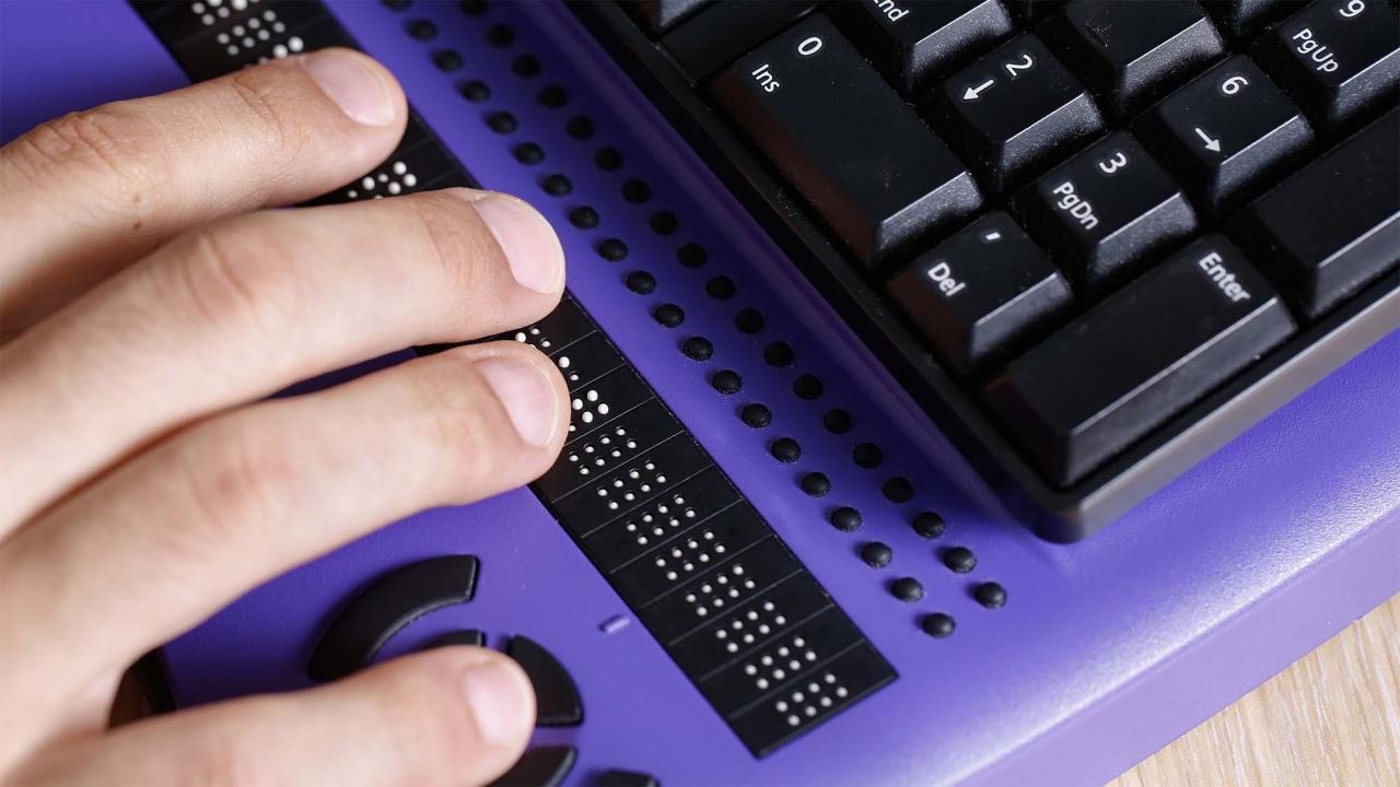 A hand rests on a purple Braille display next to a black keyboard on a wooden surface.
