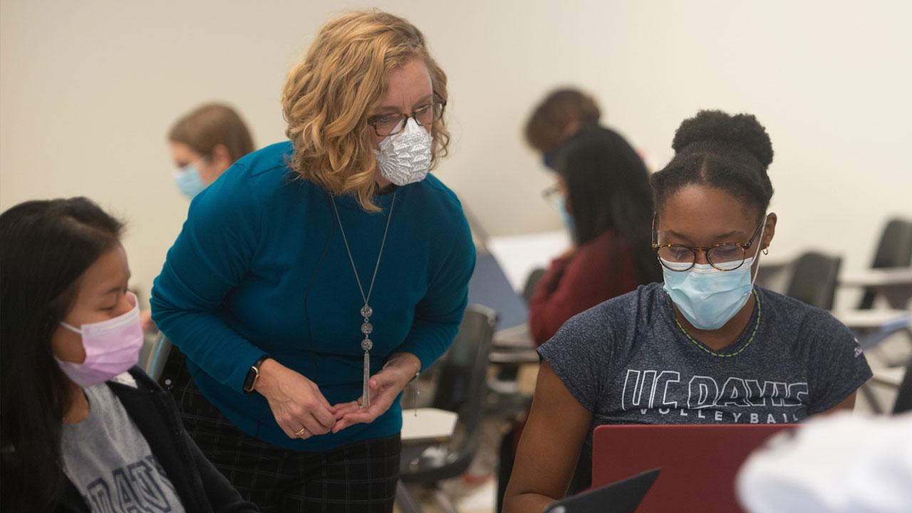 A teacher consults with some students in a classroom, all are masked