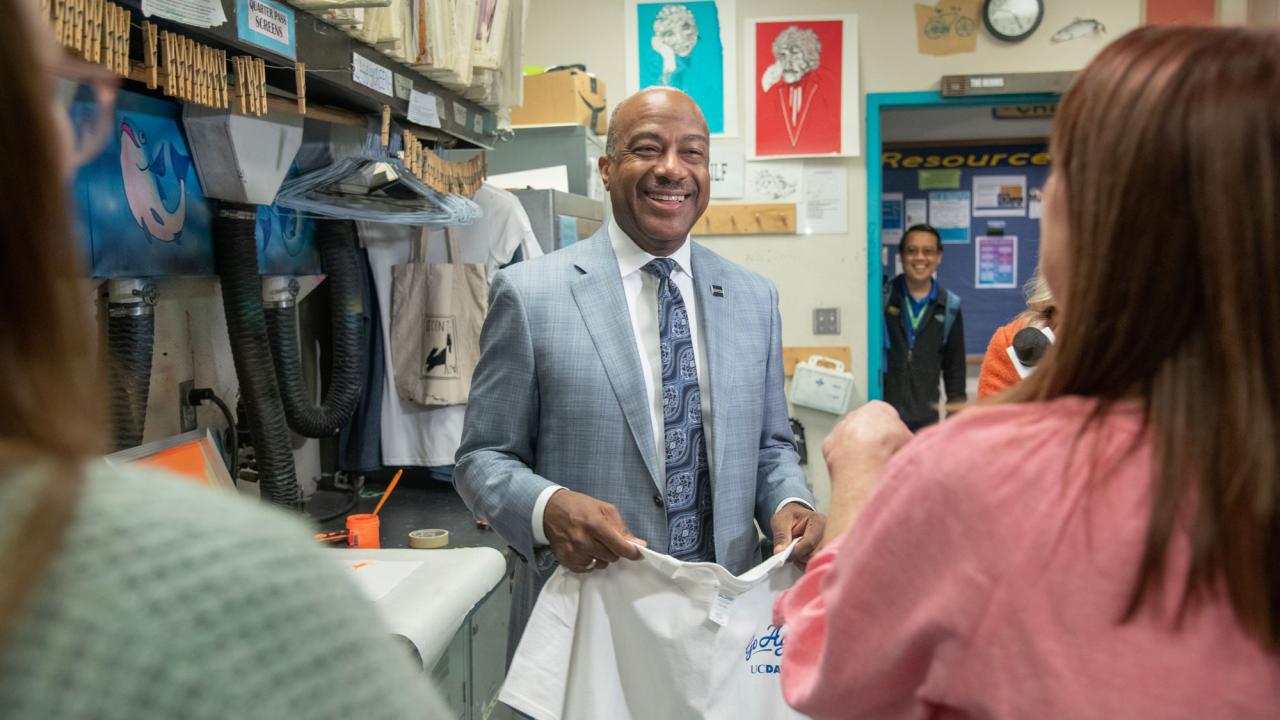 Chancellor May, in suyit and tie, smiles while holding screen-printed T-shirt and being interviewed live for morning TV show