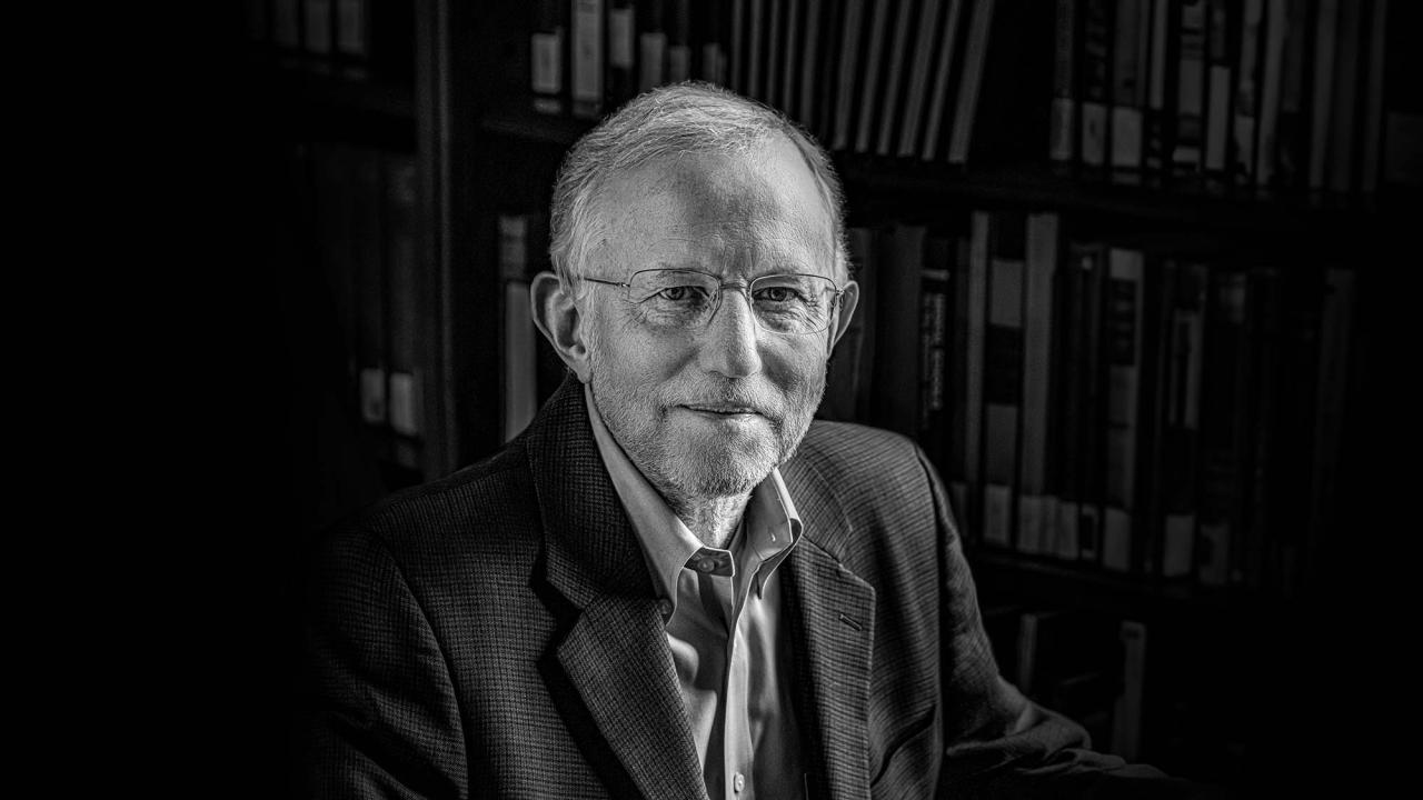 Black and white portrait of an older man with glasses. He wears a button down shirt and jacket, and smiles slightly as he looks into the camera.