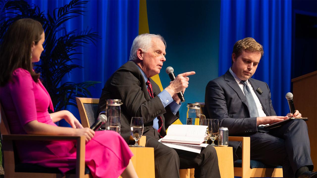 Three speakers engaged in dialogue on stage, with a blue backdrop and plants visible.