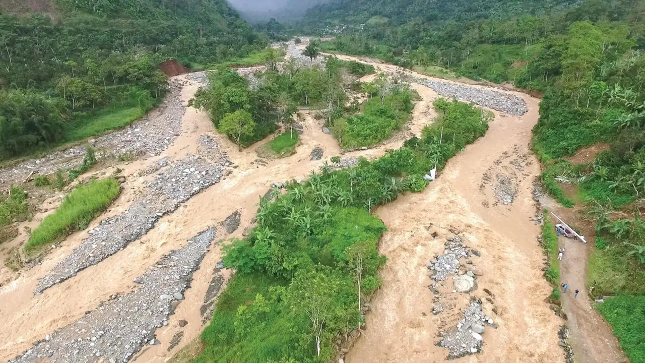 Aerial photo of a flooded river running through a narrow tropical valley. The river is muddy and fast-moving.