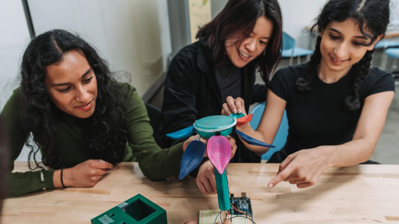 Three young women collaboratively examining a colorful, plant-like robotics project on a table.