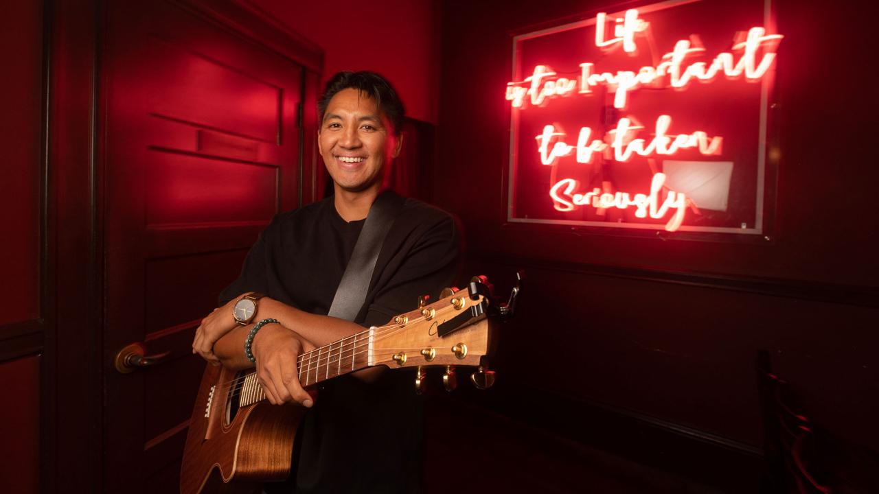 A man stands with an acoustic guitar and smiles at the camera. He appears to be backstage at a club, with a neon saying over his shoulder that read 'Life is too important to be taken seriously.'