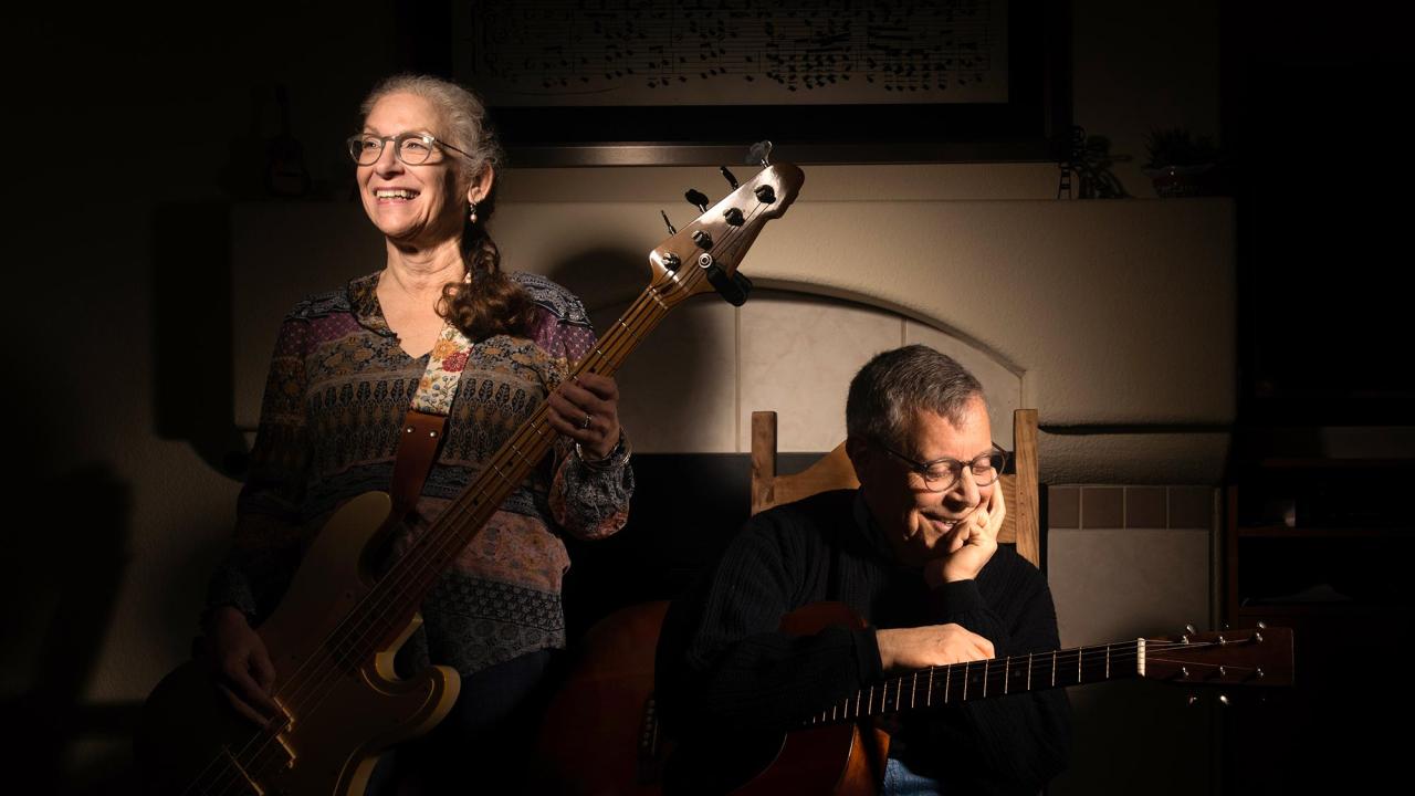 A photograph features two musicians in a warmly lit room with a fireplace in the background. The woman on the left, wearing glasses and a colorful patterned blouse, is holding a bass guitar with a leather strap, smiling brightly while looking off to the side. Her curly gray hair is pulled back. The man on the right, wearing glasses and a black sweater, sits on a wooden chair with his elbow resting on the guitar he is holding. He appears thoughtful, with a gentle smile and his hand resting on his cheek.