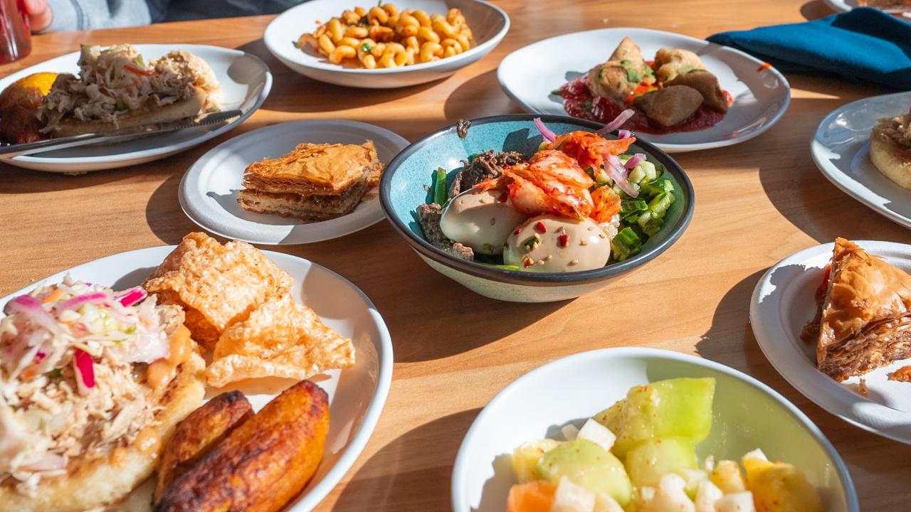 Several different plates of food sit atop an outdoor picnic table bathed in sunlight, with patrons on sitting on either side of the table. 