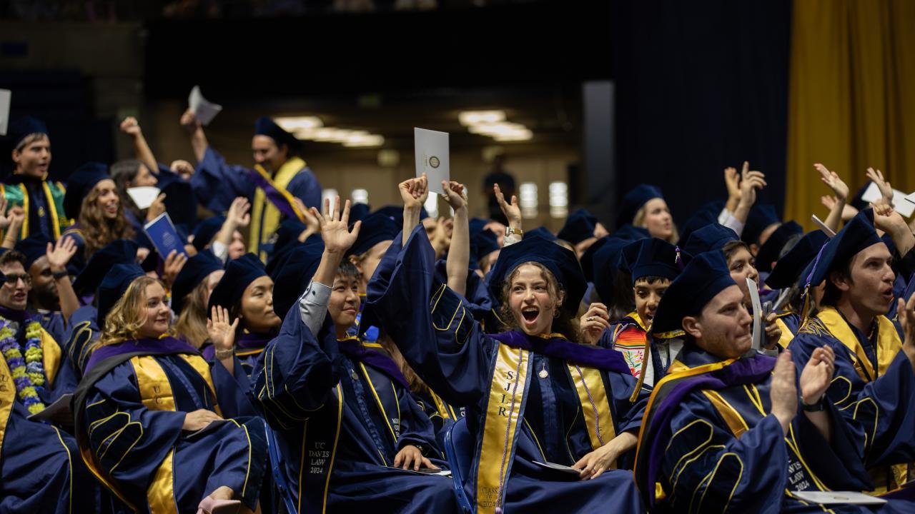 Graduates in caps and gowns celebrate joyfully, waving their hands in excitement.