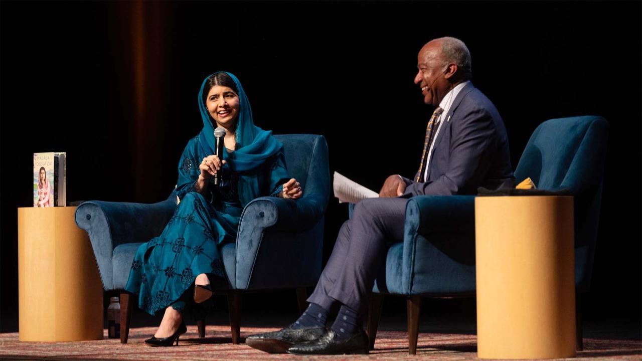 Malala Yousafzai, seated on stage, holds a microphone and speaks while Chancellor Gary S. May, seated nearby, laughs