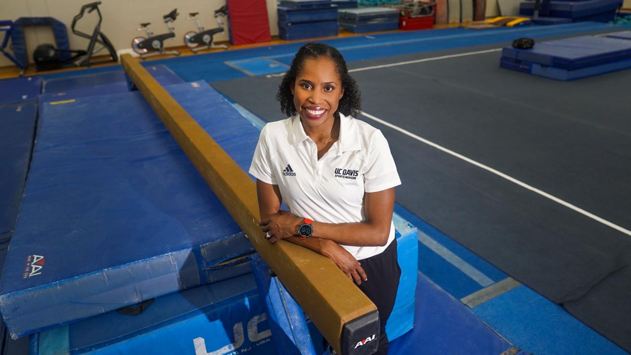 Female in white Aggie Athletics polo shirt, one arm atop balance beam in gym