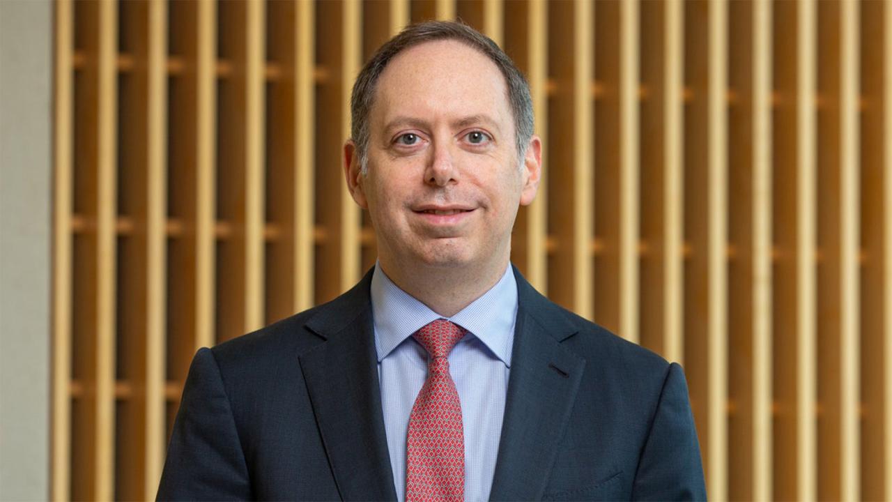 A man in a navy suit and red tie stands in front of a wood-paneled background 