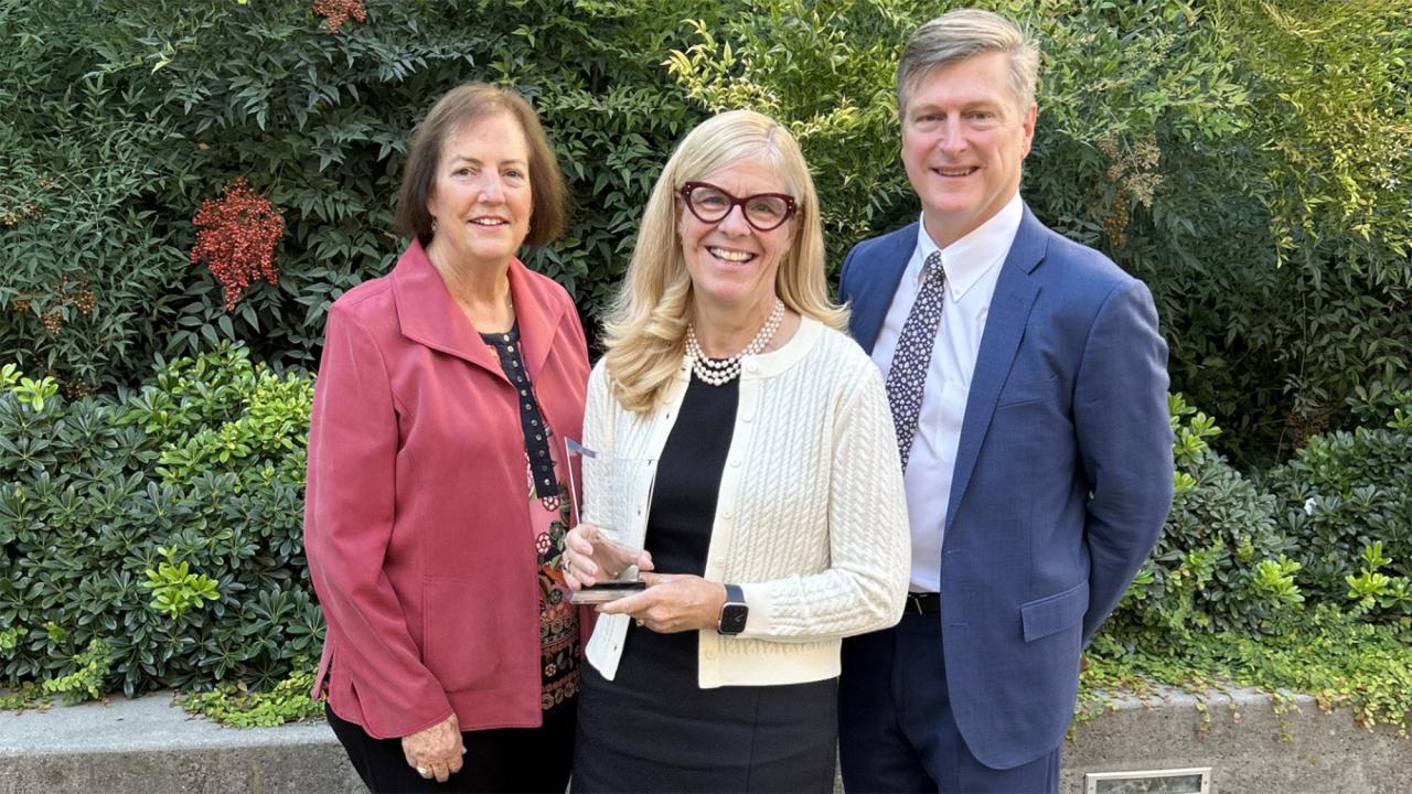 Three people pose for photo outdoors. One is holding an award.