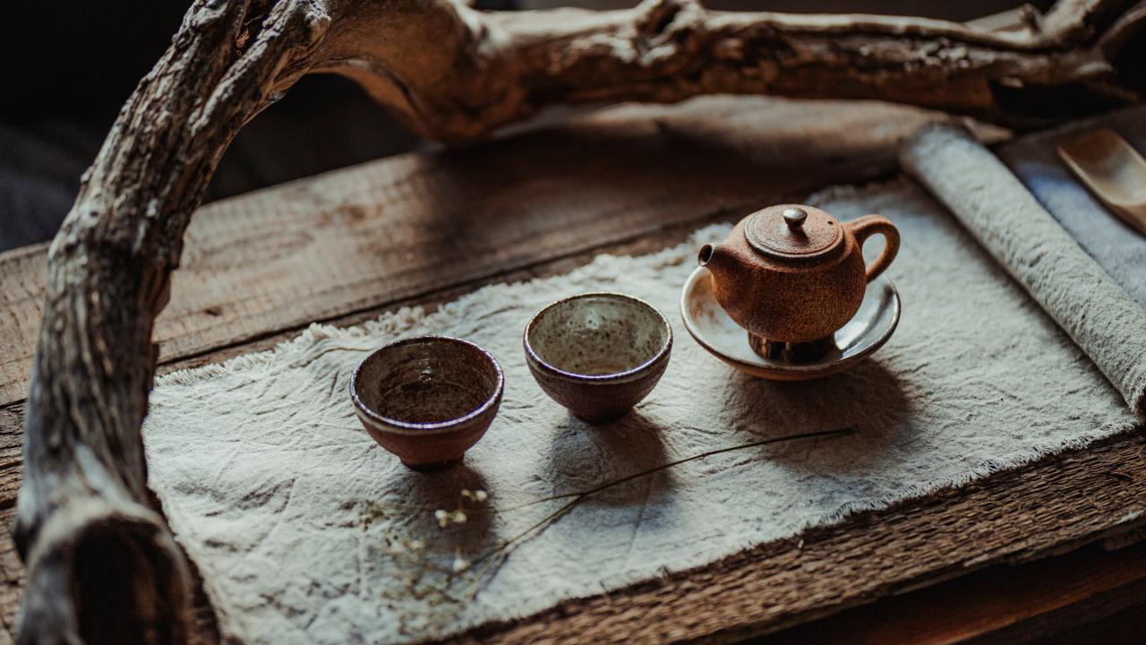 Ceramic teapot with two small cups on a textured wooden surface, framed by a branch.