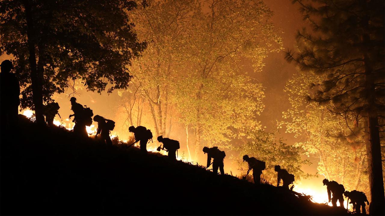 Firefighters seen in silhouette against bright flames at night.