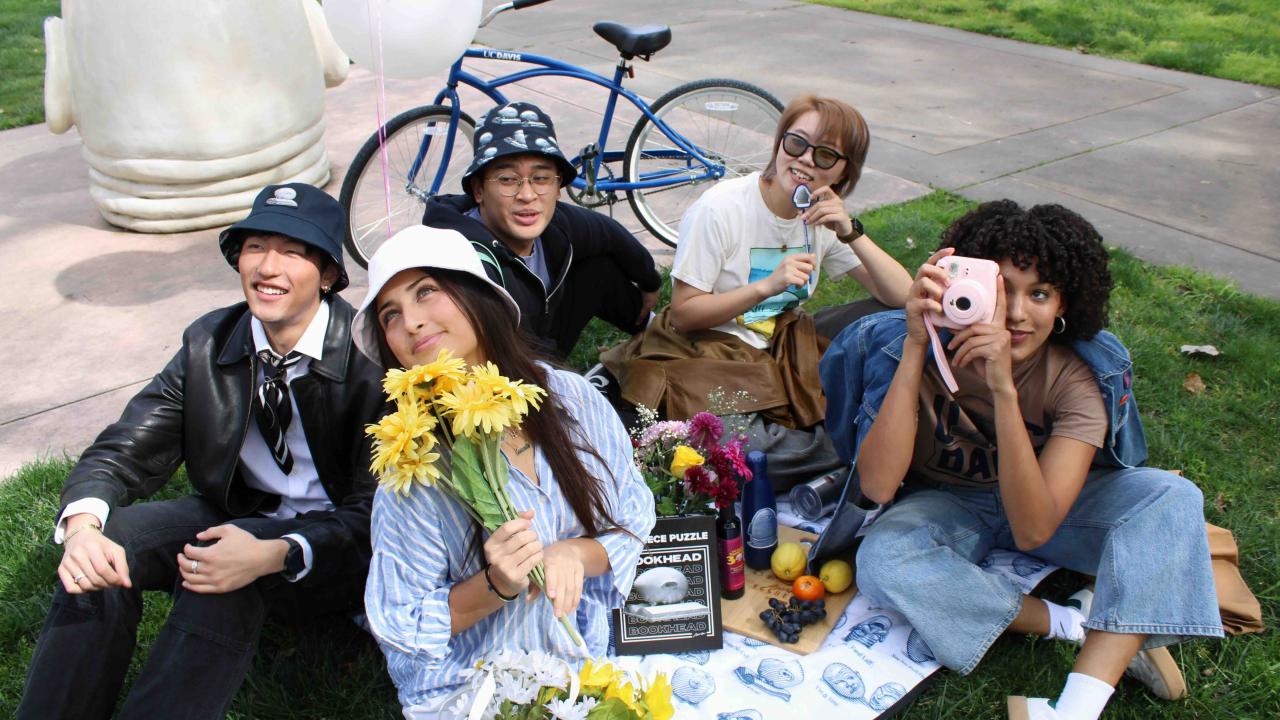 A diverse group of five Aggies enjoying a picnic on an Egghead blanked with Egghead merchandise in front of the Eye on Mrak Fatal Laff Egghead sculpture