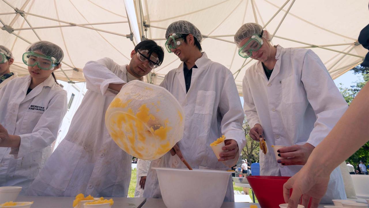 Group of people in lab coats and hair nets mix orange ice cream in large tubs outdoors
