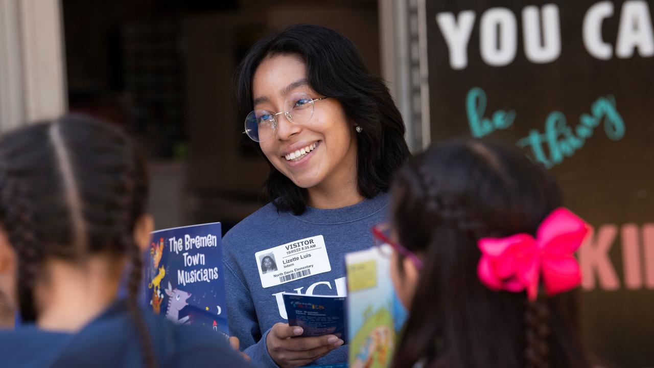 A smiling woman holding a book engages with two children outside a classroom.