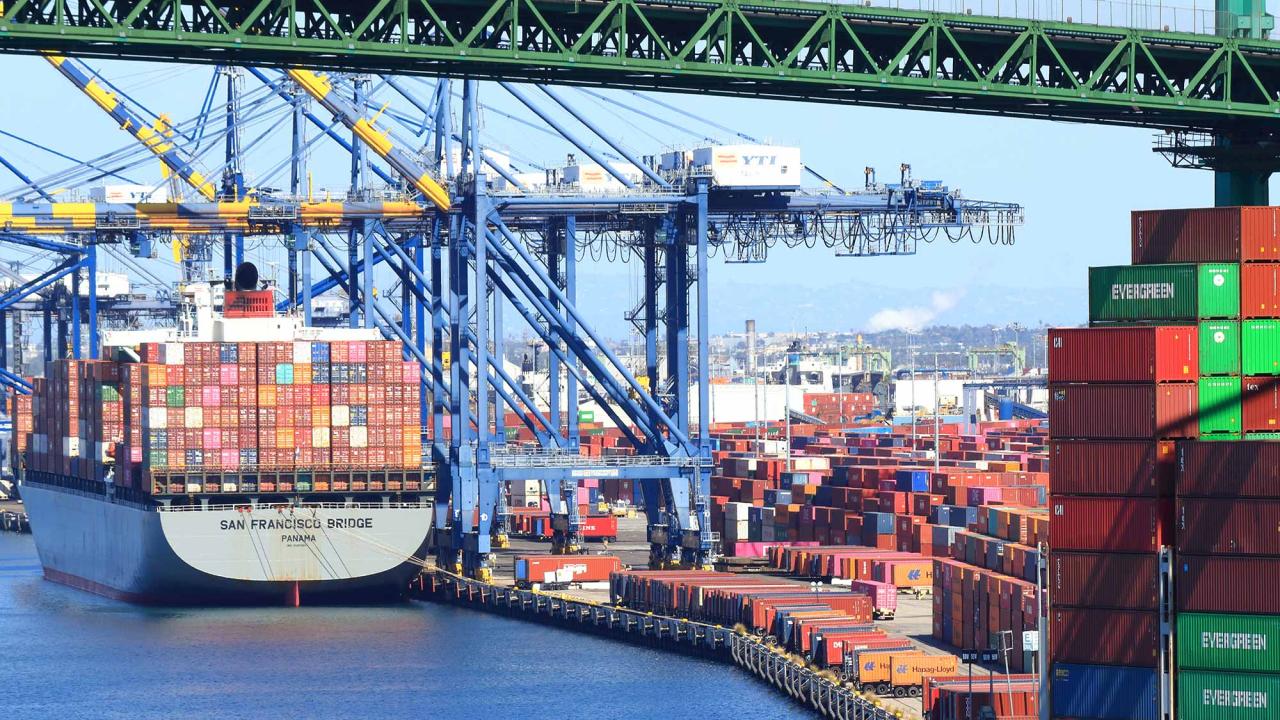 Photo of a large container ship, seen from the stern, and docked next to a large crane. Many other containers are stacked high on the dock, and the lower trusses of a green bridge can be seen spanning over the ship and port.