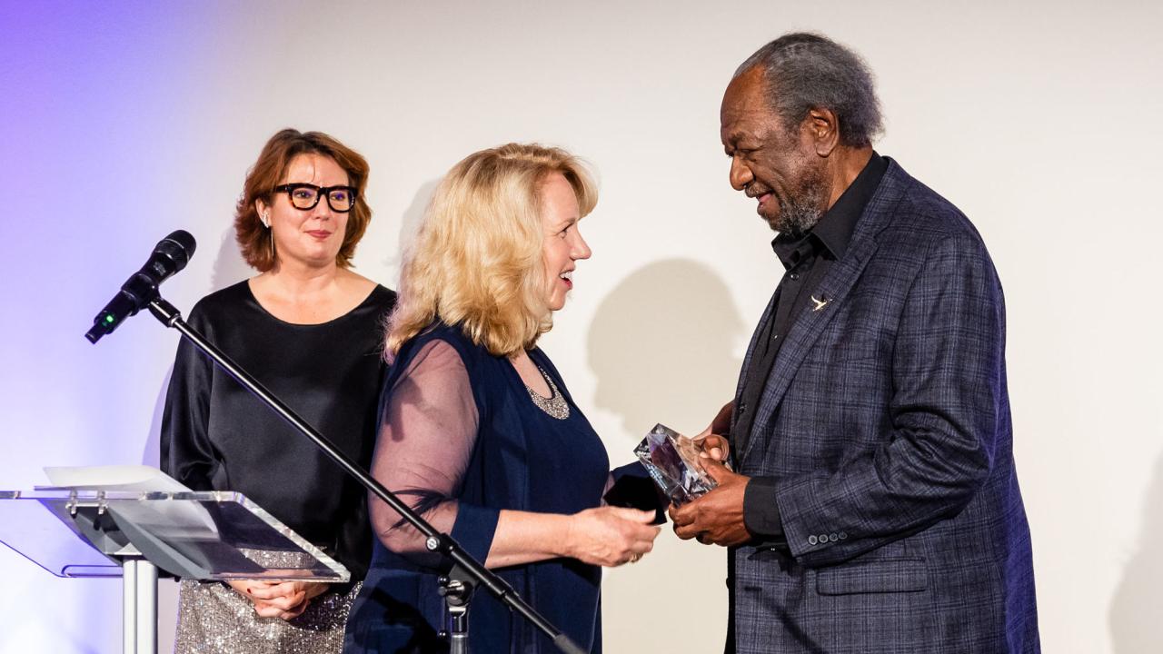 Two women present trophy to a man, on stage