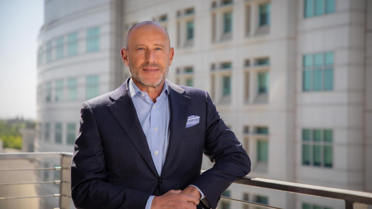 A tieless man wearing a suit stands on a balcony, juxtaposed against a building facade
