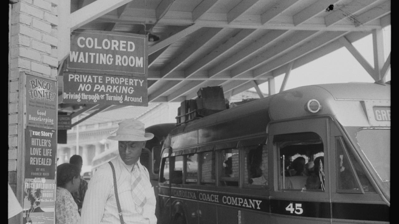 A historical black-and-white photo of a waiting area with a bus and a sign indicating "Colored Waiting Room."