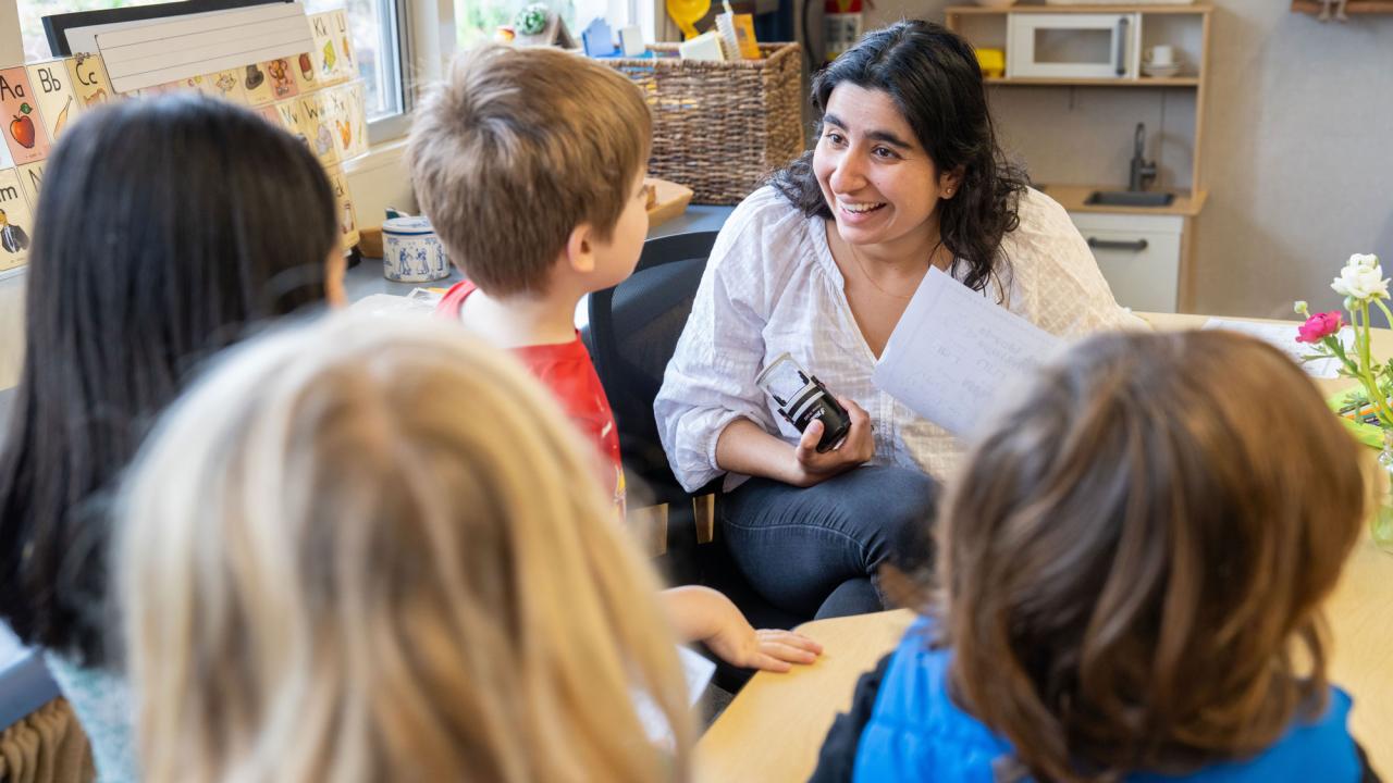 Teacher seated, with dark hair and white blouse, talks to kindergartners 