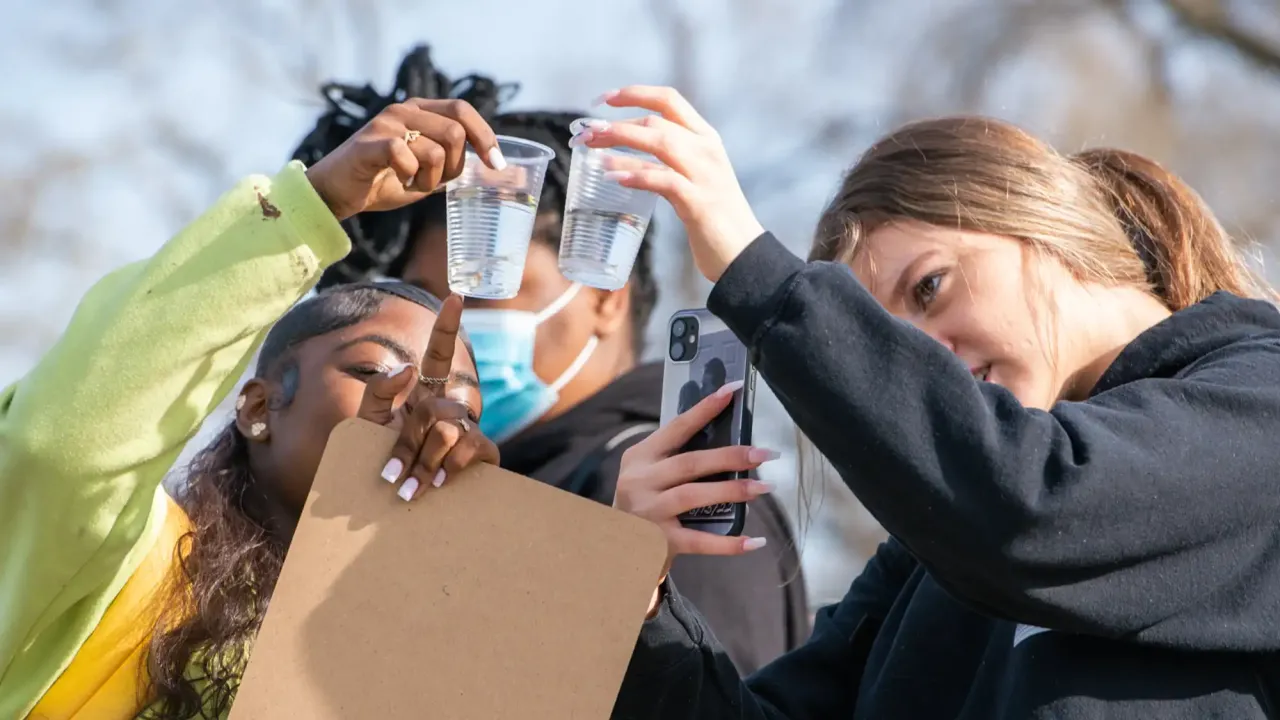 Three students closely examine clear plastic cups filled with water, holding them up to the light. One student points at a cup while holding a clipboard, another wears a face mask in the background, and a third uses a smartphone to take a photo or video. The scene takes place outdoors on a sunny day.