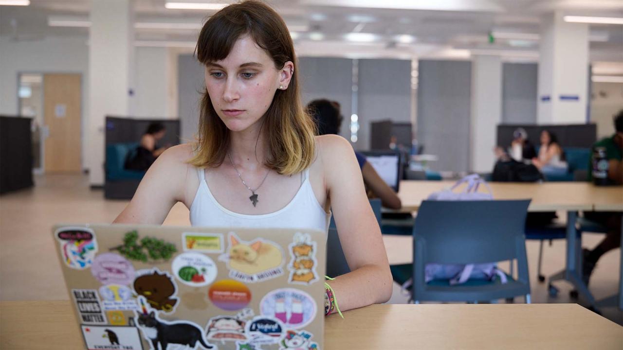Undergraduate Laila Penny sits at a table in a bright, modern study space, focused on her laptop. She has light skin, straight brown hair with blonde tips, and wears a white tank top and a lightning bolt necklace. Her laptop is covered with colorful stickers, including animals, food, and slogans like “Black Lives Matter.” Other students are visible in the background, working or socializing in small groups. 