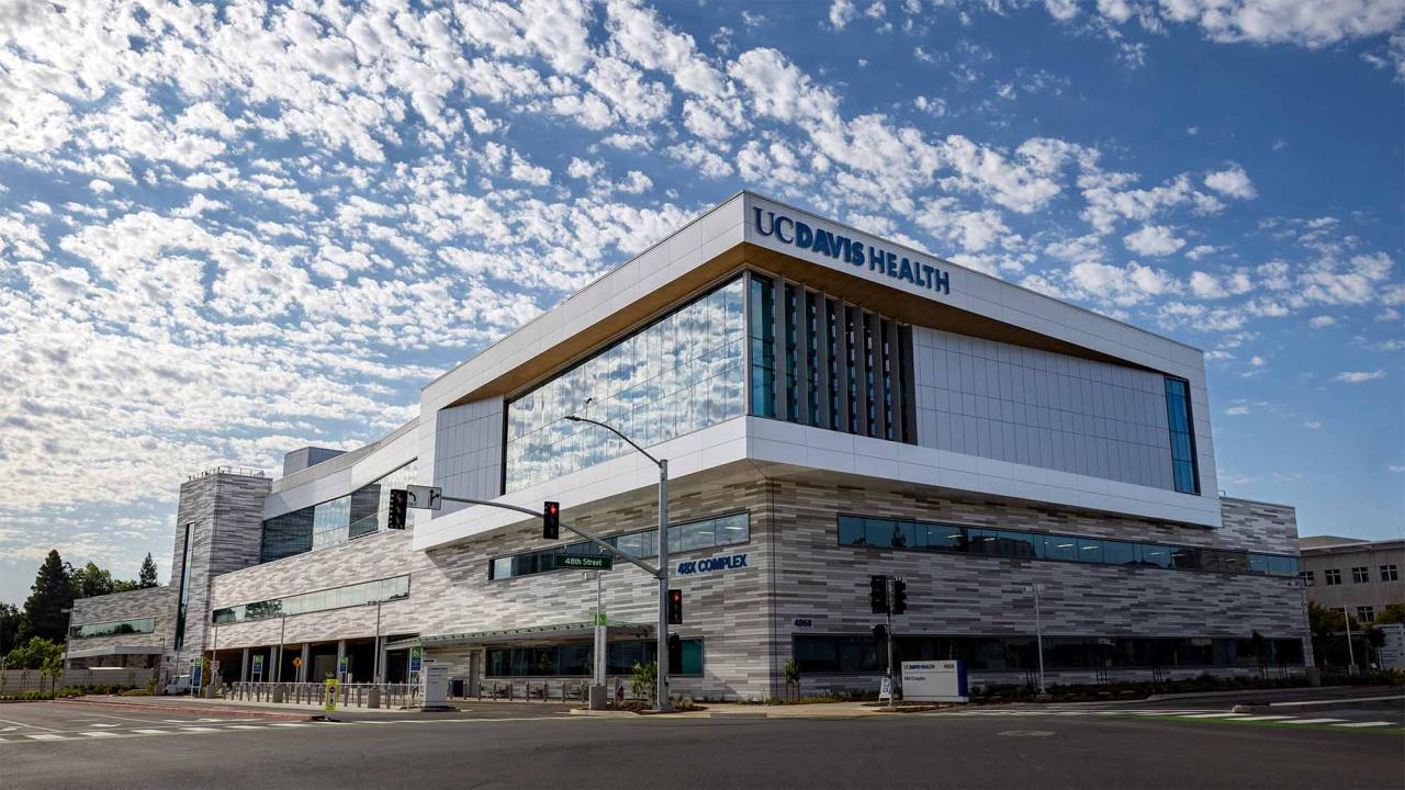 Photo shows exterior of 48X building, with UC Davis Health logo prominently visible, under bright blue sky filled with clouds.