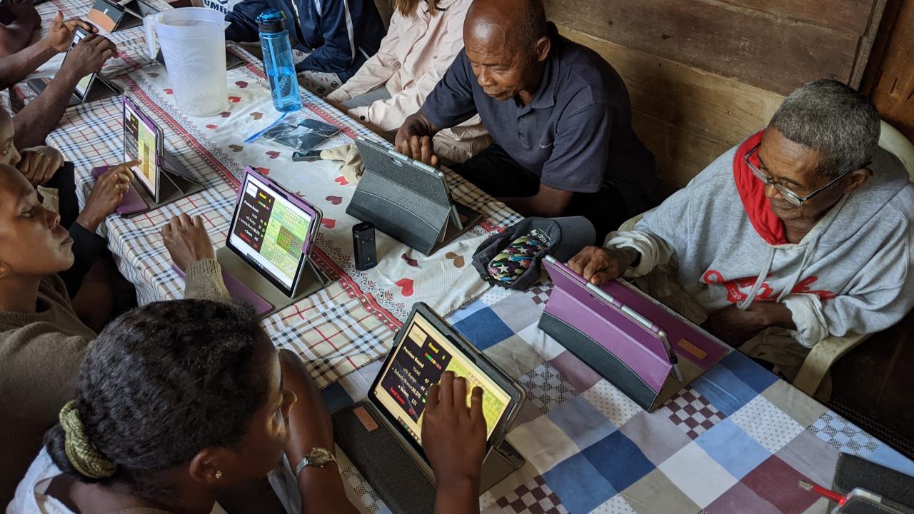 Farmers in Madagascar sit on each side of a rectangular table lined with a quilted tablecloth playing a game on digital tablets