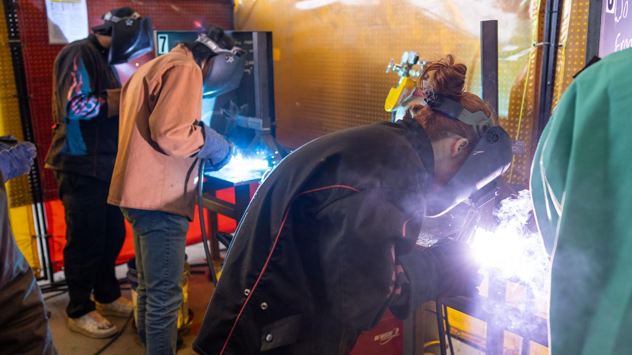 Three students in welding helmets bring bright white flame to their projects.