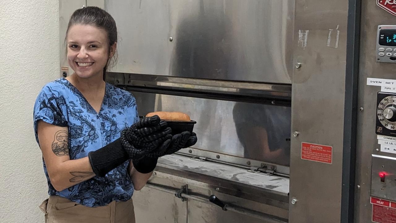 woman in blue shirt and tan pants smiles while holding loaf of freshly baked bread pulled from large metal oven