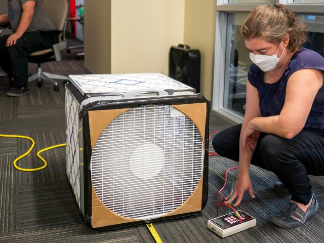A woman kneels next to a box with air filters on it to monitor the air quality of a room