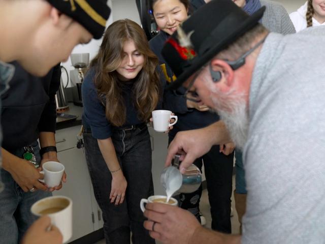 Students gather and watch with excitement as a white-bearded man in a light gray shirt, glasses, black open-ear Bluetooth headphones and a feathered black fedora pours foam into a small, white coffee mug.