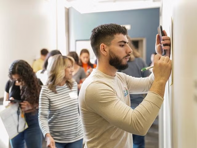 Student writing on piece of paper against a wall in classroom with other students