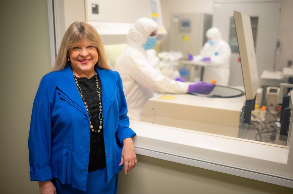 Blonde woman in blue jacket stands in front of a window looking into a biomedical lab. 