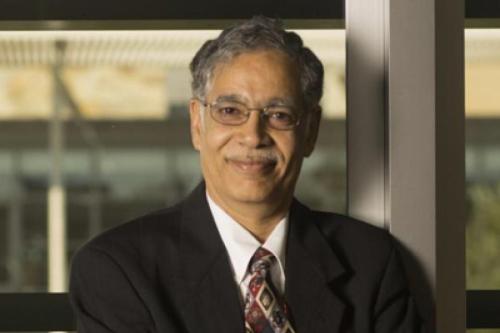 A man with grey hair smiles at the camera. He is wearing black glasses and a black suit.