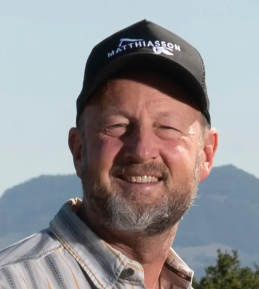 A smiling man with a beard wearing a cap and a striped shirt, standing outdoors with mountains in the background.