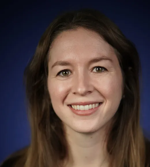 A person with long brown hair, wearing a dark green top, against a blue background.